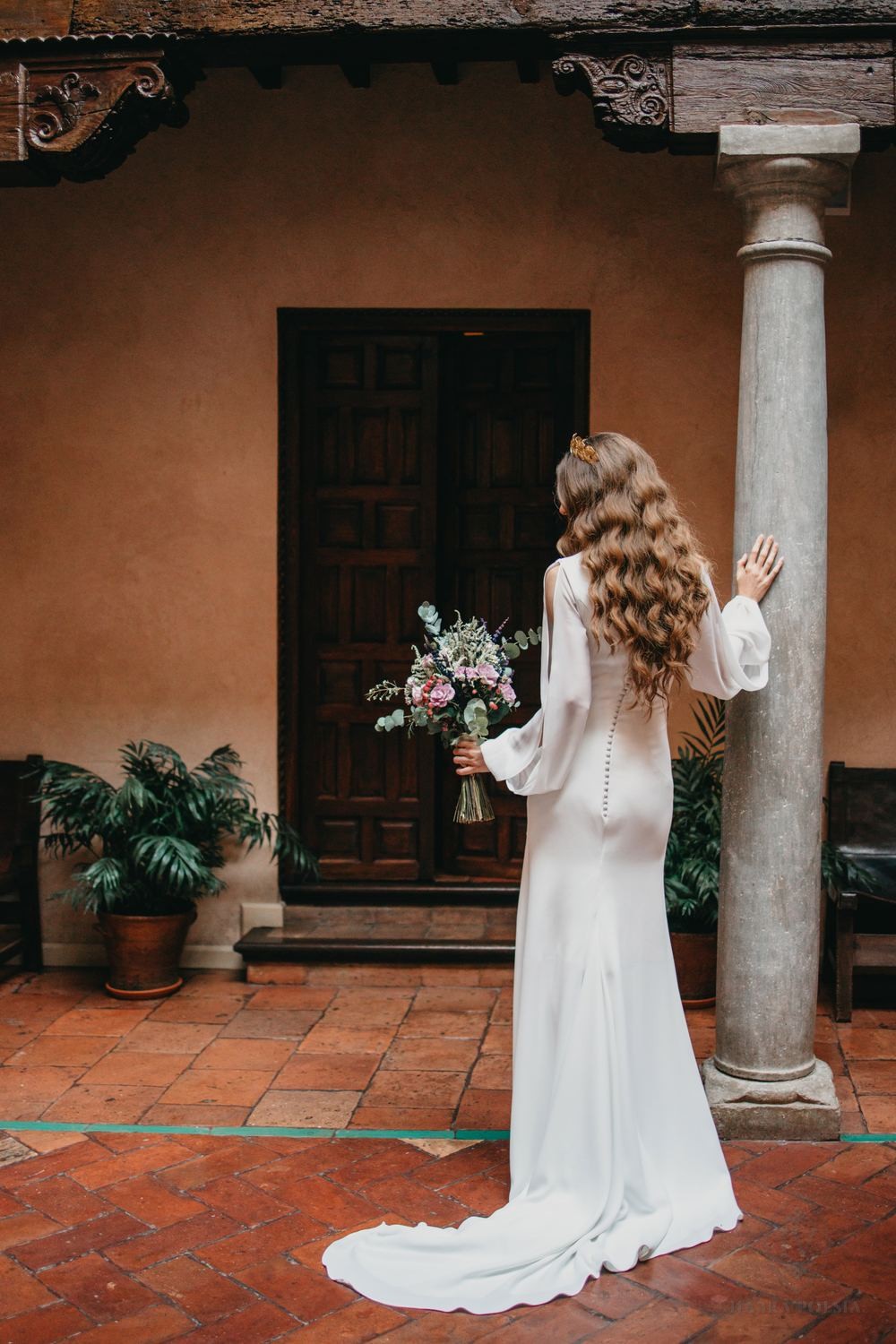 Novia con vestido de manga larga junto a una puerta de madera en un patio