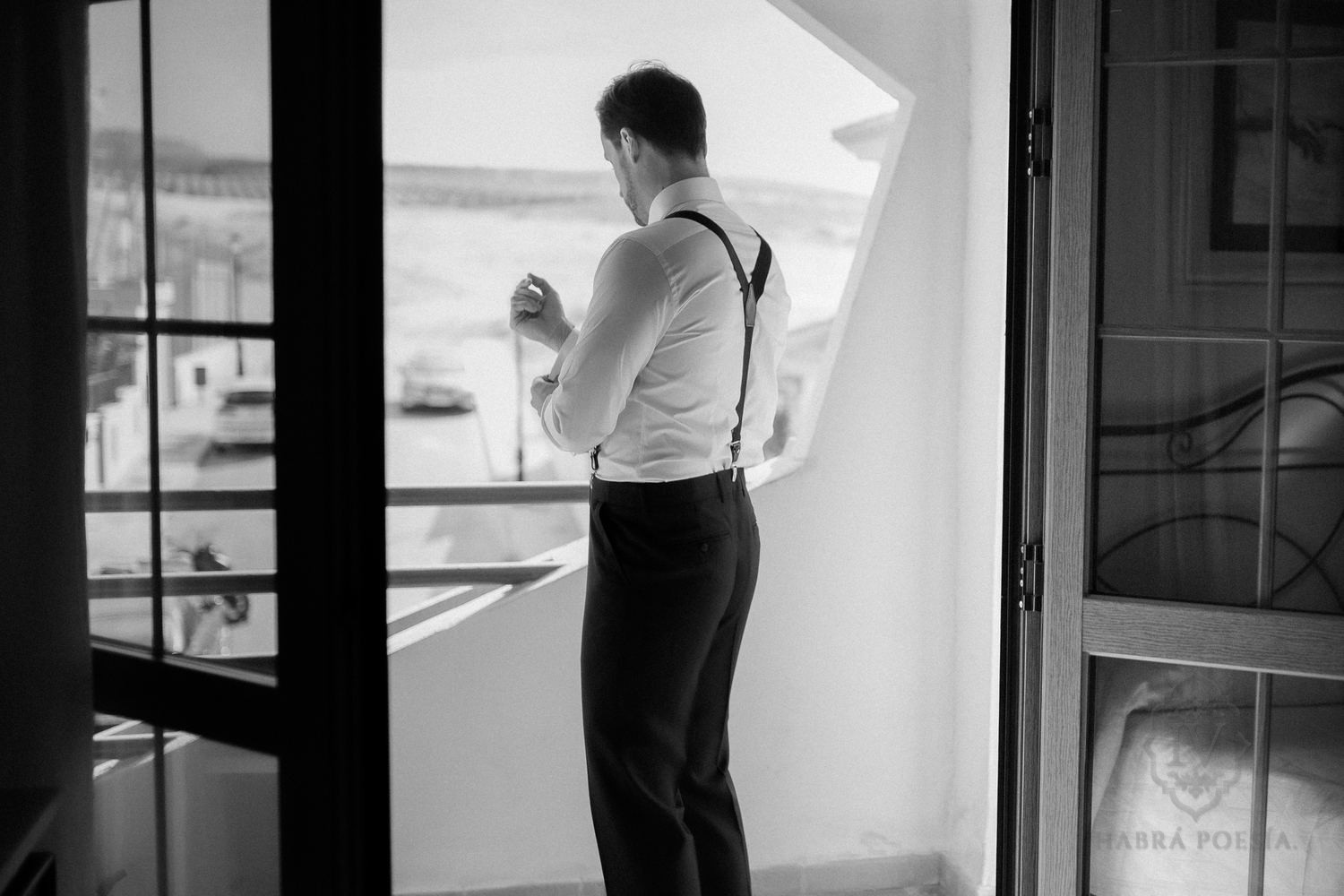 Novio con tirantes asomado a una ventana antes de la boda, en blanco y negro